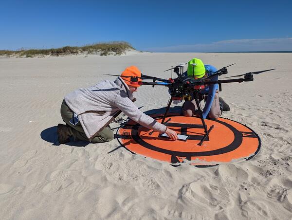 Two people kneeling on beach next to drone