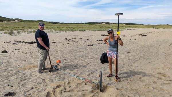 Two people installing equipment on sandy beach, vegetation in the background