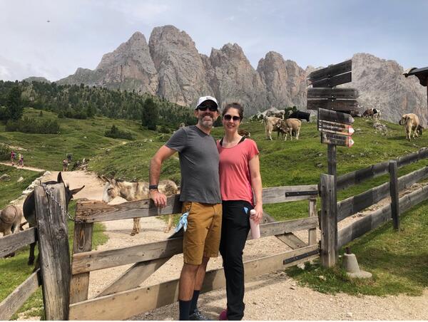 a man and woman in front of a wood fence with donkeys behind and mountains in the background