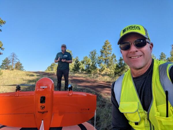 two scientists in USGS gear look into a camera, one next to a UAS and one behind it holding remote control