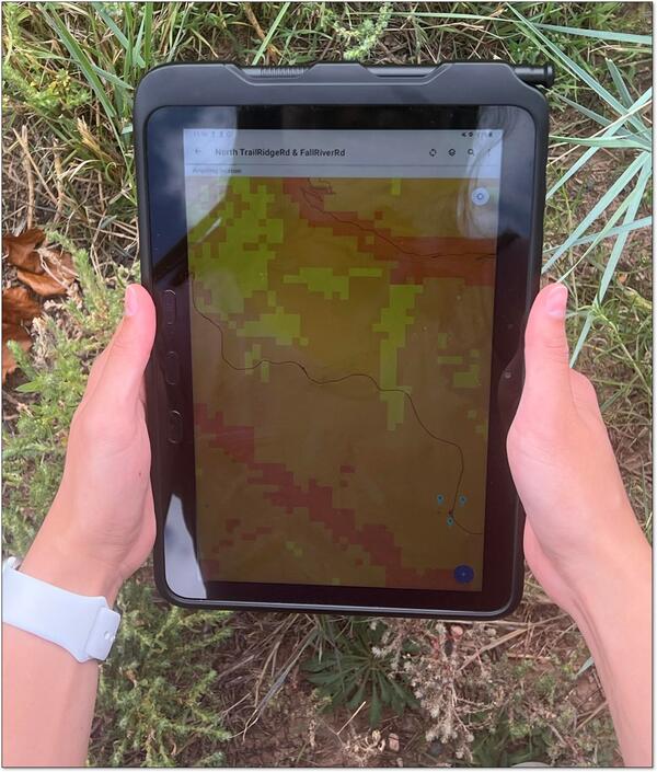 two hands hold a tablet device with grass in the background. On the tablet, a map is displayed.