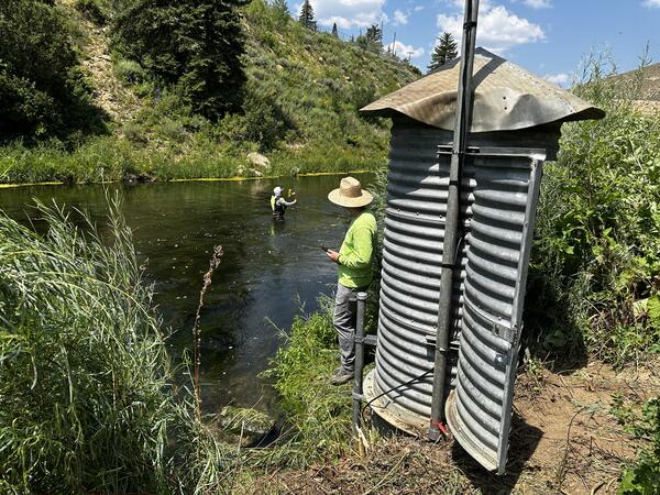 Corrugated galvanized steel gage house with tech next to it and person wading in river in background.