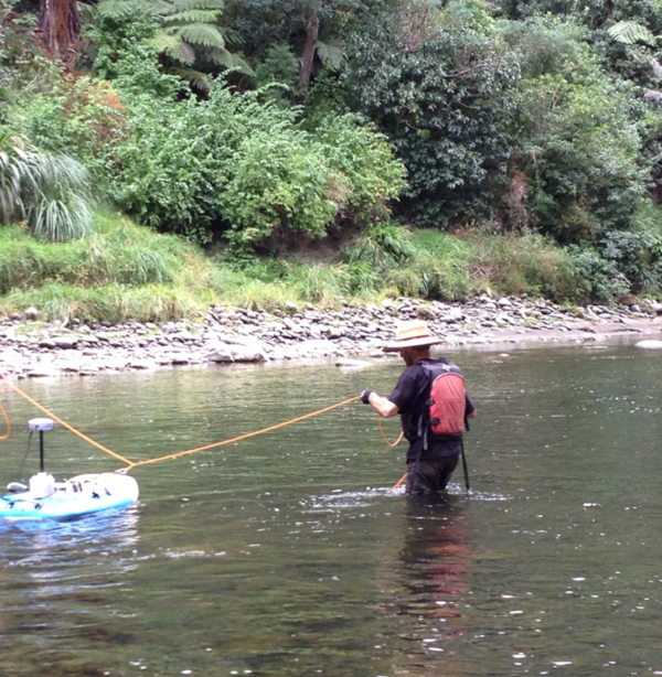 man uses a rope to pull an ADCP in a pontoon across the river. Steep, green forested river bank in distance