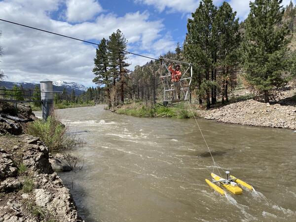 Scientist stands on platform suspended from cableway crossing a river.