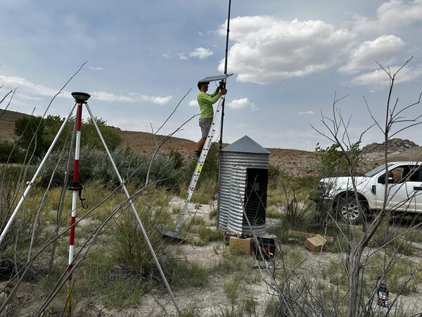 Person on ladder next to cylindrical gage house installing solar panel on pole.