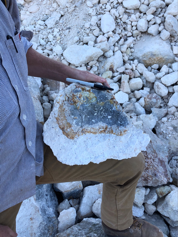 A geologist in a blue shirt and khakis supports a basketball-sized chunk of lava on his knee. A bright white "rind" makes up about half of the chunk, surrounding a core of dark gray lava speckled with white crystals. A marker rests on the chunk for a sense of scale, and similar white chunks of lava make up the background of the photo. 