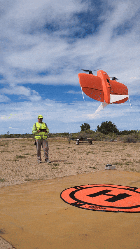 A man in neon yellow gear holds a remote control, an orange drone rises up and lands on a helipad in front of the man