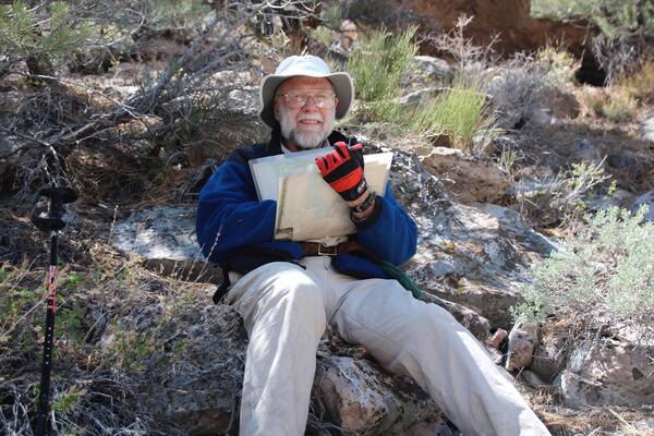 A man with white hair and a beard, wearing khakis, a blue shirt, and a fisherman's hat, reclines on a rock outcrop. He is holding a bundle of geologic maps and smiling.