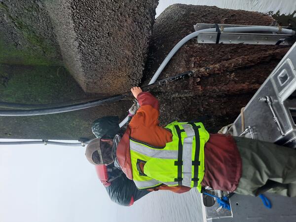 Two men stand on the edge of a boat right up against a concrete bridge pier covered in barnacles 