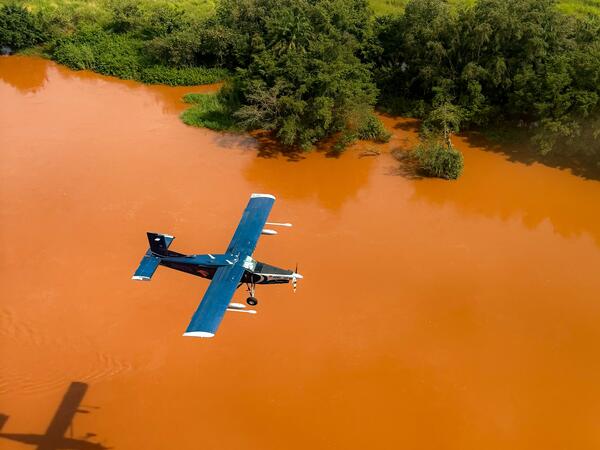 Aerial view of a dark blue single-engine plane flying over a river or floodplain stained bright orange-red