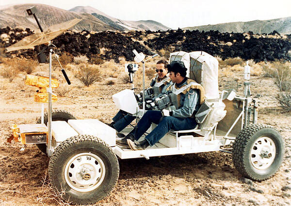 Two white men wearing jeans, button-down shirts, and bulky harnesses with huge rectangular backpacks sit on the frame of a stripped-down white motorized buggy-like vehicle. Their harnesses have two old-fashioned video cameras pointing forward. On the front of the vehicle, a large metallic mesh radio antenna dish is raised on a pole. In the background, mounds of dark, jagged, black lava rock sit in front of a skyline composed of several rounded red hills of volcanic scoria. 