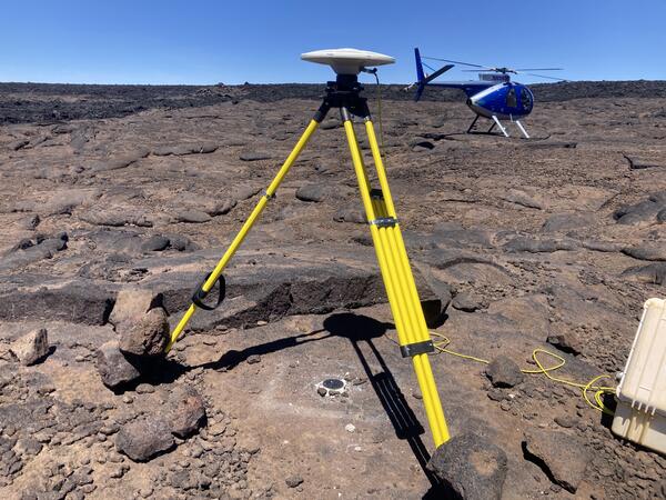 Color photograph of tripod on lava flow with helicopter in the background