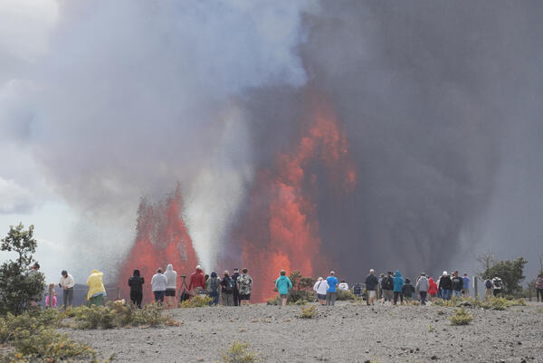 Color photograph of people standing on the rim of a crater with lava fountains in the background