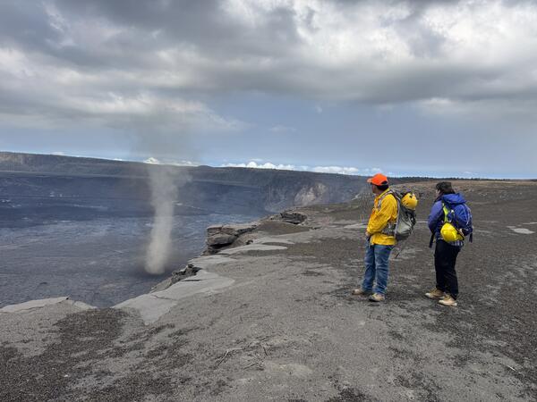 Color photograph of scientists on the edge of a volcanic crater with a whirlwind in it