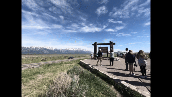 timelapse photography of people walking up to an entrance sign, clouds moving above mountains, cars driving by