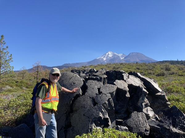 Man standing next to rock with hat and high-vis vest. Snow covered mountain in background.
