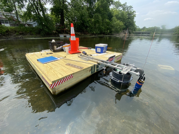 Orange traffic cone, sample storage box, and a notebook sit on a wooden platform in the river with the Bacteria sampler.