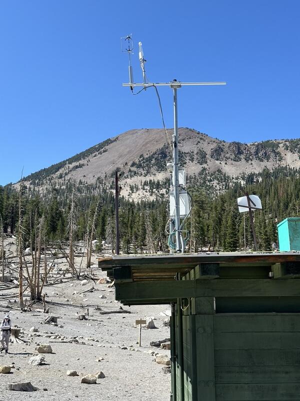 A tall white metal tower with scientific instruments stands on a rooftop of a small building in a mountainous area with scattered rocks and sparse vegetation. In the background, Mammoth Mountain rises as a forested peak under a clear blue sky. The monitoring equipment includes sensors mounted at the top of a 6-meter-high mast for measuring volcanic CO₂ emissions.