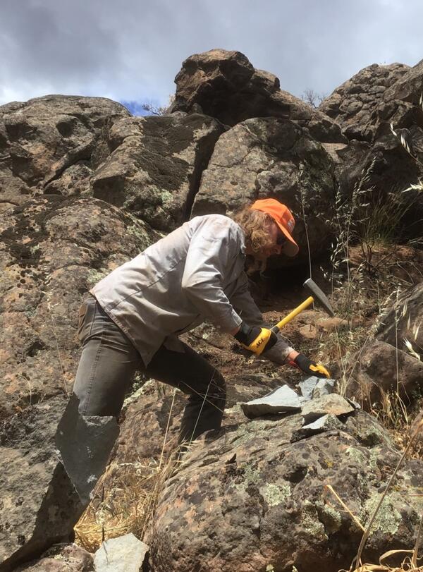 A geologist wearing a blue button-down shirt, khaki pants, and an orange ball cap uses a rock hammer to break smaller chunks off the large, weathered, brownish boulders that surround him. The interior of the chunks is revealed to be a pale, speckled gray.