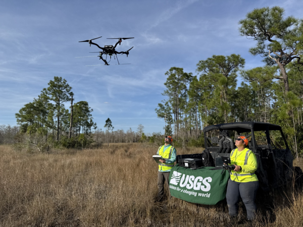 two people in yellow vests lean against an OHV, look up at drone flying above them, grasses and trees in background