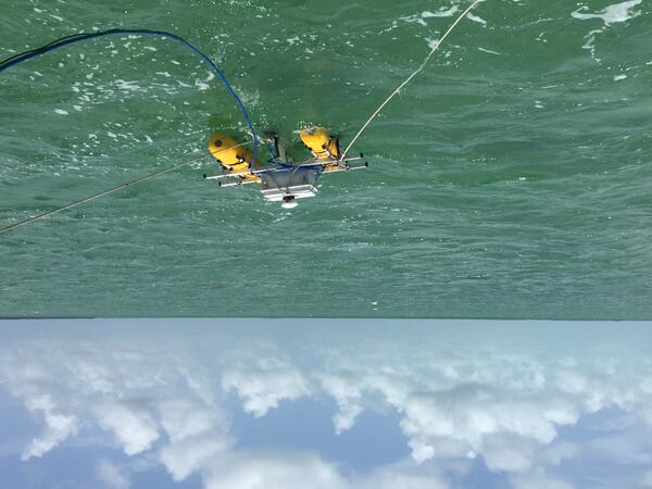 Scientific equipment mounted on two yellow pontoons floating on the surface of green water under a blue sky
