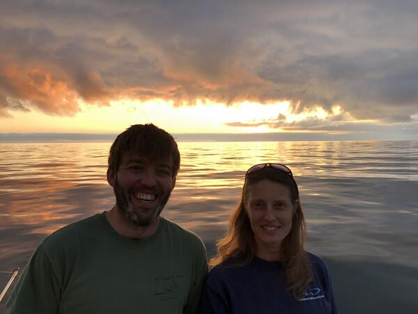 A man and woman stand together smiling with the sun setting over the water behind them.