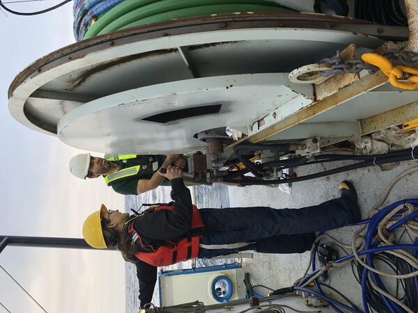 Two people stand on the deck of a boat near a large cable winch.