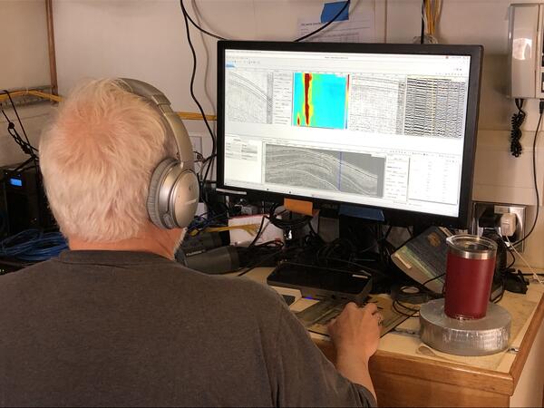 A man sits at a table, looking at a computer monitor, he is wearing headphones.