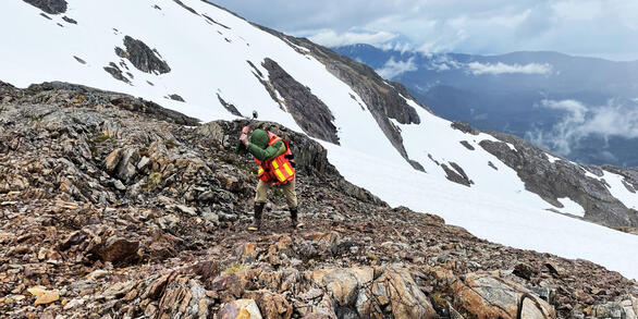 USGS geologist breaks mineralized rocks for examination and sample collection. Snow on slopes behind geologist.