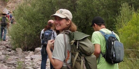 Image of geneticist Caren Goldberg in the field. She is wearing a backpack and stands in front of some vegetation