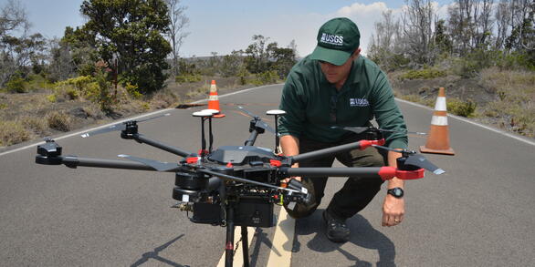a man in dark green USGS gear kneels next to a large black drone in the middle of a road, cones and trees in the background