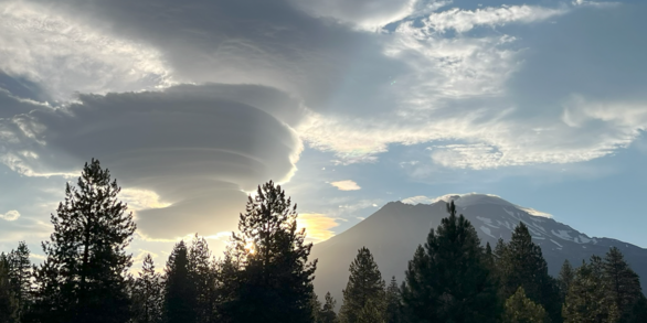 A photograph taken on the forested lower slopes of a mountain looks toward a snowy peak. A stack of saucer-shaped clouds to the left of the mountain are illuminated by the rays of the sun as they peek over the slopes. In the foreground, pine trees are thrown into stark contrast by the dramatic sky above.