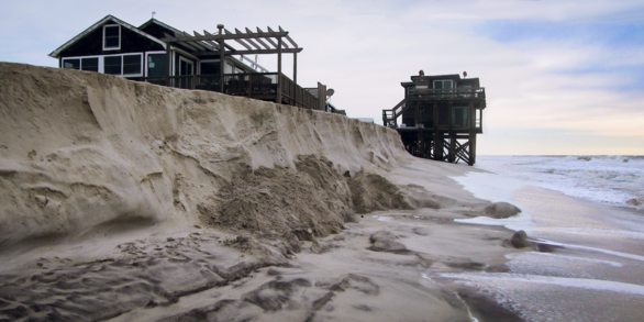 a sandy coastline with heavily eroded dunes with two houses on stilts nearby