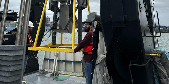 Two scientists guide sampling equipment attached to large crane, on to deck of research vessel