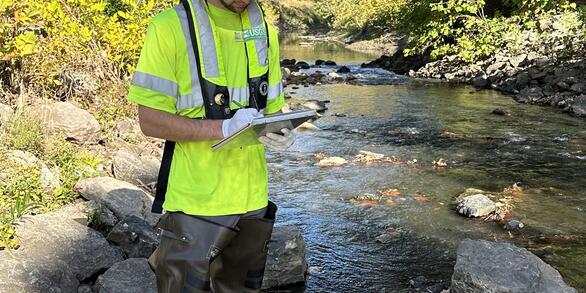 Scientist records conditions of river while standing next to it with a highway overpass in background