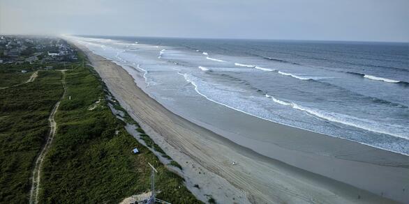 A beach with vegetated dunes and ocean waves approaching the shoreline. Houses are seen on the island in the distance.