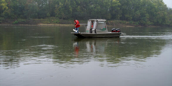 USGS scientist stands on a boat deck, lowering a water-quality sampler into a river