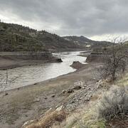 Brown river water flows by muddy banks with course grained sediment. Columns of basalt in right. Cloudy skies.