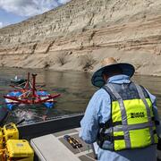 Scientist wearing safety gear sits in boat, operating equipment. Towed equipment can be seen on water behind boat.