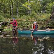A man in a red vest stands in knee-deep water in a river with a handheld device while another man paddles a canoe.
