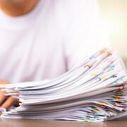 a man in a white shirt signs a paper with stack of paper clipped papers next to his hands