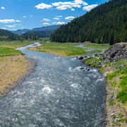 A blue strip of river runs through grassy, rocky fields. Steep hills with pine trees line both sides of the image