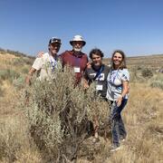 Four people stand behind a large sagebrush plant with shrubby landscape and blue sky in the background.