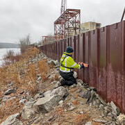 A man in a neon yellow jacket measures a high water mark on a wall with a river in the background.