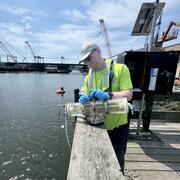A man in a cap, neon yellow shirt, and blue gloves holds a Van Dorn sampler on a dock with a streamgage and bridge behind.