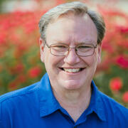 A man in a blue collared shirt wearing glasses and seated in front of red flowers.