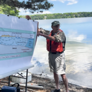 A man in a baseball cap and orange lifejacket stands in front of a lake and points to a sign.