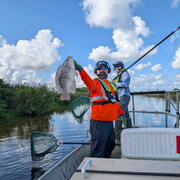 two people in life vests stand in a boat - one holds a fish while the other holds a dipnet