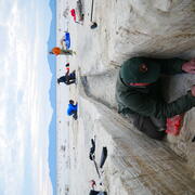 Person works with fine tools in a sandy trench, with others working in the background and mountains in the distance
