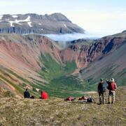 Two people standing by a ledge facing and gesturing away towards mountains in the distance; packs and two people on ground